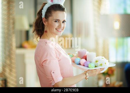 Lächelnde junge Hausfrau in einer rosafarbenen Bluse und österliche Hünenohren, die am sonnigen Frühlingstag einen Teller mit osterdekoration im modernen Zuhause hält. Stockfoto