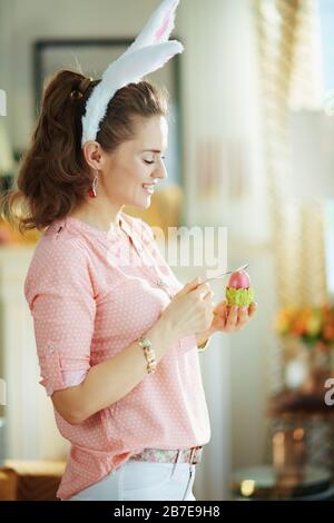 Fröhliche, moderne 40 Jahre alte Hausfrau in einer rosafarbenen Bluse und osterbrötchen, die rotes osterei in grünem Eierbecher mit Löffel im modernen Haus in su essen Stockfoto