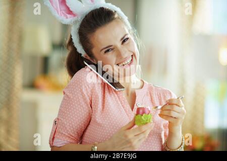 Fröhliche moderne Frau in einer rosafarbenen Bluse und osterbüschen mit einem Handy beim Essen von rotem osterei in grünem Eierbecher mit Löffel zu Hause Stockfoto