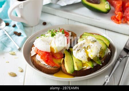 Frühstück am Morgen. Pochiertes, mit Ei gekochtes Sandwich mit Avocado, Lachs und Kaffee auf einem Holztisch. Stockfoto