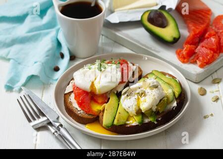 Frühstück am Morgen. Pochiertes, mit Ei gekochtes Sandwich mit Avocado, Lachs und Kaffee auf einem Holztisch. Stockfoto