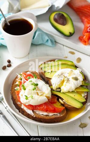 Frühstück am Morgen. Pochiertes, mit Ei gekochtes Sandwich mit Avocado, Lachs und Kaffee auf einem Holztisch. Stockfoto