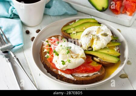 Frühstück am Morgen. Pochiertes, mit Ei gekochtes Sandwich mit Avocado, Lachs und Kaffee auf einem Holztisch. Stockfoto