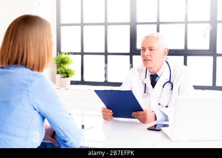 Schuss eines männlichen Arztes mittleren Alters, der mit seinem Patienten spricht, während er am Büroschreibtisch im Arztraum sitzt. Stockfoto