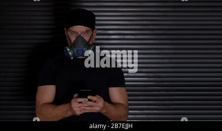 Ein Mann mit einer Atemmaske, der auf eine schwarz gekleidete Kamera blickt, mit einem Handy in den Händen auf dunklem Hintergrund. Coronavirus Konzept Stockfoto