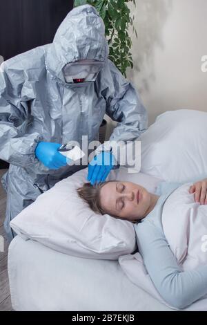 Die Person, die einen Schutzanzug, eine Maske und eine Schutzbrille trägt, misst die Temperatur einer kranken Frau, die in ihrem Bett liegt. Bekämpfung der Epidemie. Stockfoto