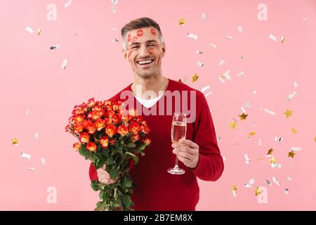 Bild eines positiven, glücklichen, reifen Mannes mit Lippenstift auf dem Gesicht isoliert über dem rosafarbenen Wandhintergrund in rotem Pullover, der Blumen und Champagner über dem Confet hält Stockfoto