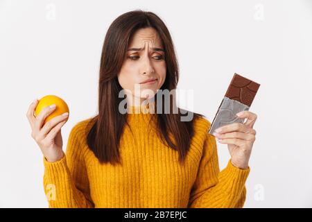 Bild einer zweifelhaften Frau, die Erwachsene brünette, zögert, während sie Orange und Schokoladenriegel isoliert über weißem Hintergrund hält Stockfoto