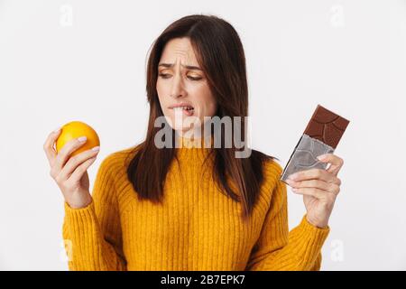 Bild einer zweifelhaften Frau, die Erwachsene brünette, zögert, während sie Orange und Schokoladenriegel isoliert über weißem Hintergrund hält Stockfoto