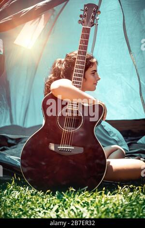 Teenager-Mädchen, die gerne Urlaub im Sommercamp verbringen, hübsches Kind, das mit einer Gitarre in der Nähe des Zelts sitzt, Lied singt und Spaß hat Stockfoto