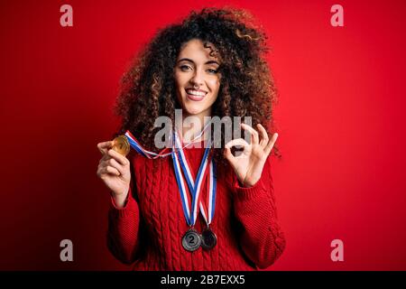 Junge schöne und erfolgreiche Frau mit lockigen Haaren und durchstechenden Medaillen, die mit den Fingern in Ordnung sind, hervorragendes Symbol Stockfoto