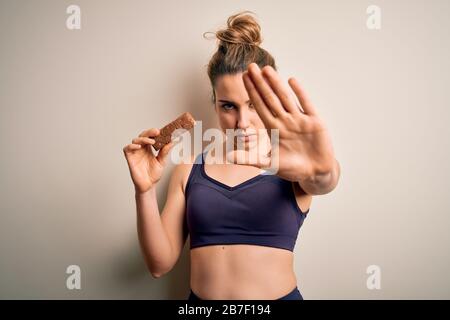 Junge schöne blonde Sportlerin mit Sportswear und energischer Eiweißbar mit offener Hand und Stoppschild mit ernstem und selbstbewussten Expressi Stockfoto