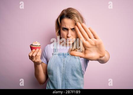 Junge schöne blonde Frau Eatimg Schokoladenkuchen über isoliertem rosafarbenem Hintergrund mit offenem Handzeichen mit ernsthaftem und selbstbewusstem Expressi Stockfoto