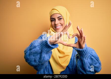 Junge schöne brünette muslimische Frau, die arabischen Hijab über isoliertem gelbem Hintergrund trägt, lächelnd in der Liebe, die mit den Händen eine Form des Herzsymbols tut. Romantisch Stockfoto