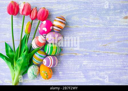 Ostereier und schöne rosafarbene Tulpen in verschiedenen Designs und Farben werden auf einem pastellvioletten Holzhintergrund mit Kopierraum für den Fall während EA platziert Stockfoto