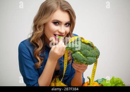 Süße Frau an einem Tisch, die einen Brokkoli auf dem Hintergrund von Obst und Gemüse hält Stockfoto