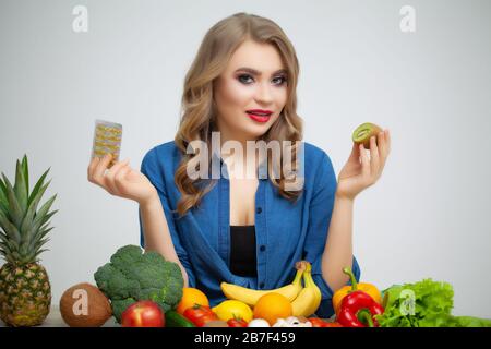 Frau an einem Tisch mit Kiwi und Pillen auf dem Hintergrund von Obst und Gemüse. Stockfoto