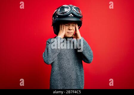 Junge, kleine kaukasische Kinder, die einen Motorradhelm mit Vintage-Biker tragen und über rotem Hintergrund googles, das an Kopfschmerzen leidet, verzweifelt und gestresst beca Stockfoto