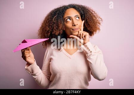 Junge afro-amerikanische Frau, die Papierflieger über rosafarbenem Hintergrund hält, ernstes Gesicht, das über die Frage nachdenkt, sehr verwirrte Idee Stockfoto