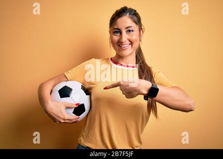 Schöne brünette Spielerin, die mit Fußball über gelbem Hintergrund Fußball spielt, sehr glücklich, mit Hand und Finger zu zeigen Stockfoto