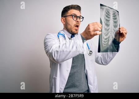 Junger Arzt, der einen medizinischen Mantel trägt, der die Röntgendiagnostik des Brustkorbs über einem isolierten Hintergrund betrachtet, erschreckt in einem Schock mit einem überraschenden Gesicht, Angst und Erregung Stockfoto