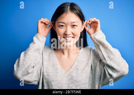 Junge, schöne asianerin, die einen lässigen Pullover trägt, der über blauem, isoliertem Hintergrund Steht, Lächelnde Ohren mit Fingern zieht, lustige Geste. Audition p Stockfoto