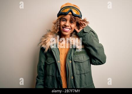 Junge afroamerikanische Skifahrerin mit lockigen Haaren, die Schneesportkleidung trägt und eine Skibrille, die mit der Hand über dem Ohr lächelt, um Gerüchten oder zuzuhören Stockfoto