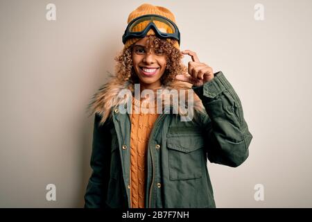 Junge afroamerikanische Skifahrerin mit lockigen Haaren, die Schneesportkleidung und eine Skibrille trägt, lächelnd und selbstbewusst mit der Hand in kleiner Größe gestikulieren Stockfoto