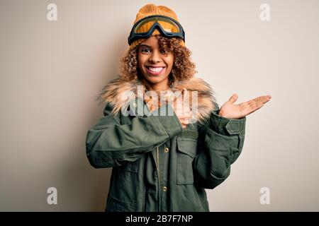 Junge afroamerikanische Skifahrerin mit lockigen Haaren, die Schneesportkleidung und eine Skibrille Trägt, Die Handfläche zeigt und mit Daumen nach oben geht, schmilzelt Stockfoto