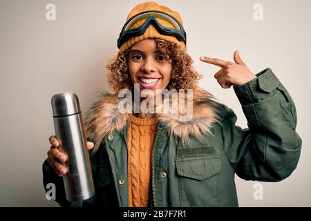 Junge afroamerikanische Skifahrerin, die eine Skibrille trägt und Thermo mit Kaffee trinkt, mit einem überraschenden Gesicht, das mit dem Finger auf sich zeigt Stockfoto