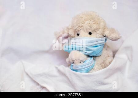 Kleine Spielzeugschafmutter mit Lamm in Händen mit Phonendoskop liegt im Bett. Spielzeug in medizinischer Schutzmaske bei Kinderspielen. Coronovirus, Quarantäne, Stockfoto
