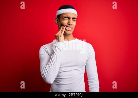 Junge, gutaussehende afroamerikanische Sportlerin, die Sportswear über rotem Hintergrund trägt und den Mund mit der Hand berührt, mit schmerzhafter Ausdrucksweise wegen Zahnschmerzen Stockfoto
