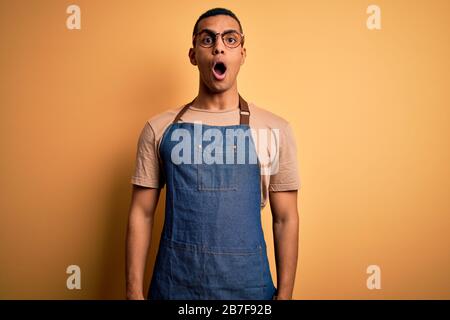 Junger gutaussehender afroamerikanischer Ladenbesitzer, der Schürze über gelbem Hintergrund trägt, fürchtet sich und schockiert mit Überraschungsausdruck, Angst und aufgeregtem Gesicht Stockfoto