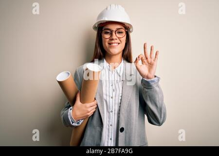 Junge, schöne Architektin, die einen Sicherheitshelm und eine Brille trägt, die Blaupausen hält, die mit den Fingern in Ordnung sind, ein hervorragendes Symbol Stockfoto