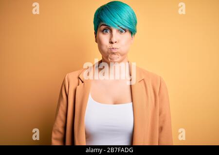 Junge schöne Frau mit blauem Modehaar mit legerer Jacke über gelben Hintergrund auffälligen Wangen mit lustigem Gesicht. Mund mit Luft aufgeblasen, cra Stockfoto