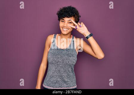 Junge schöne afro-amerikanische afro-Sportlerin, die Sport macht und Sportswear Trägt, Mit den Fingern über dem Gesicht ein Symbol für Frieden tut und fröhlichen Showin lächelt Stockfoto