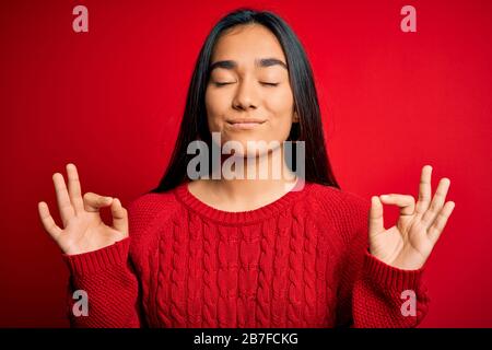 Junge schöne asiatische Frau mit legeren Pullover über isoliertem rotem Hintergrund entspannen Sie sich und lächeln Sie mit geschlossenen Augen, die Meditationsgeste w ausführen Stockfoto