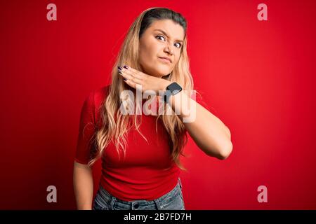 Junge, schöne blonde Frau mit legeren T-Shirts, die mit der Hand als Messer über isoliertem rotem Hintergrund stehen, bedrohen Aggressionen mit Stockfoto