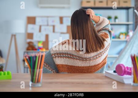 Junge, schöne Lehrerin, die Pullover und Brille auf dem Schreibtisch im Kindergarten Trägt, Denkt Rückwärts über Zweifel mit der Hand am Kopf nach Stockfoto