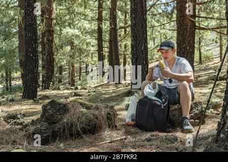 Der junge Reisende in einem cp mit Rucksack und Wanderstab sitzt auf einem Stein und hat ein Mittagessen und ruht. Kiefernwald in der Nähe des Vulkans Arenas Negras. Teneras Stockfoto