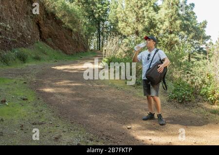 Junge Reisende in einer Kappe mit Rucksackhaltestellen auf der örtlichen Straße halten eine Plastikflasche Wasser und ruht. Kiefernwald in der Nähe des Vulkans Arenas Negr Stockfoto