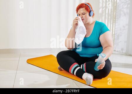 Junge übergewichtige Frau, die nach dem Training im Gesundheitsclub Gesicht wischt und frisches Wasser trinkt Stockfoto