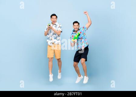 Smilng glückliche asiatische Männerfreunde, die mit Wasserpistolen spielen und in blau isoliert auf den Hintergrund für das Songkran Festival in Thailand und Südostasien springen Stockfoto