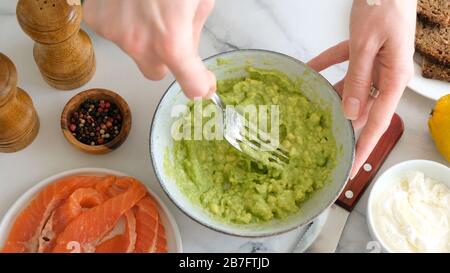 Avocado mit einer Gabel in der Schüssel auf dem marmornen Tischhintergrund verzurren. Kochen gesundes Essen mexikanische Guacamole-Avocado-Sauce, sauberes Esskonzept Stockfoto