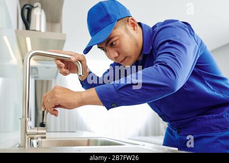 Seriöser junger Installateur in blauer Uniform, der Hahn im Küchenwaschbecken installiert Stockfoto