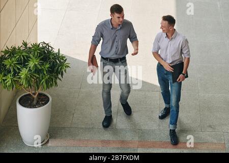 Fröhliche junge Geschäftsleute, die vor dem Bürogebäude spazieren und Nachrichten diskutieren Stockfoto
