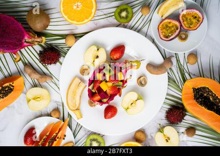 Obere Ansicht der Platten mit geschnittenen Früchten. Auswahl an verschiedenen exotischen Früchten mit tropischen Blättern auf Marmorgrund. Köstlicher Snack, Vitaminheilt Stockfoto