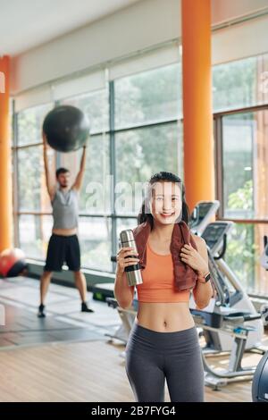 Schöne, fröhliche junge Vietnamesin mit Handtuch und kleinen Thermoskannen, die nach dem Training am Morgen im Fitnessstudio stehen Stockfoto