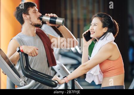 Fröhliche junge Vietnamesin telefoniert beim Fahrradfahren, ihr Trainer steht in der Nähe und trinkt frisches Wasser Stockfoto