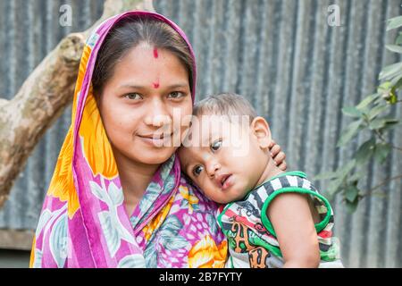 Eine lächelnde Mutter mit Zinnoberrot auf der Stirn steht mit ihrem Baby vor ihrem Haus im Bezirk Bagerhat im Süden von Bangladesch. Stockfoto